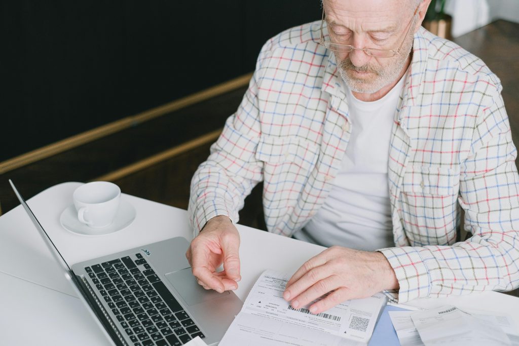 elderly man in glasses reviewing financial documents with a laptop open on his desk